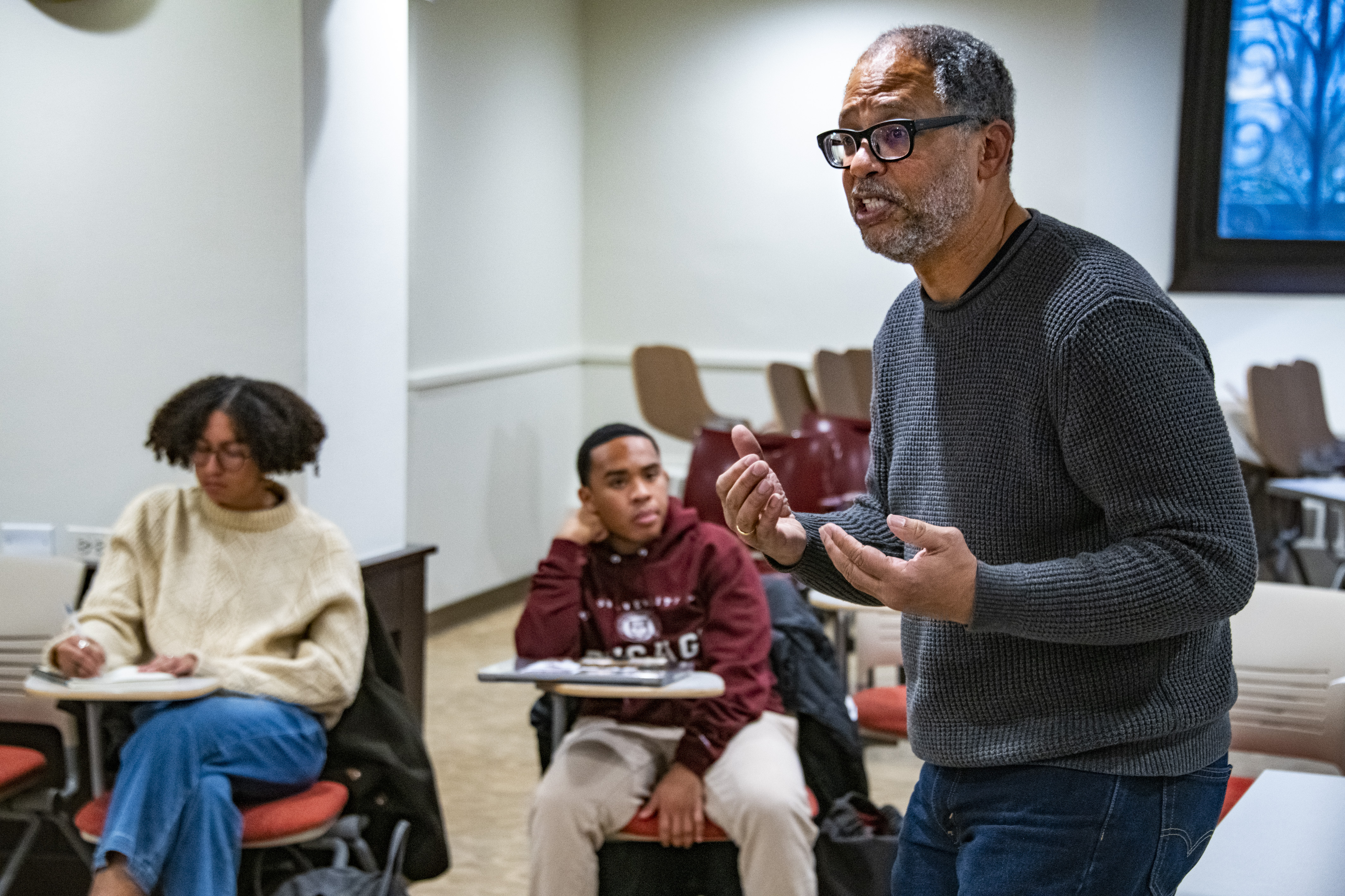 Professor lectures to room full of students, with two students behind him listening.
