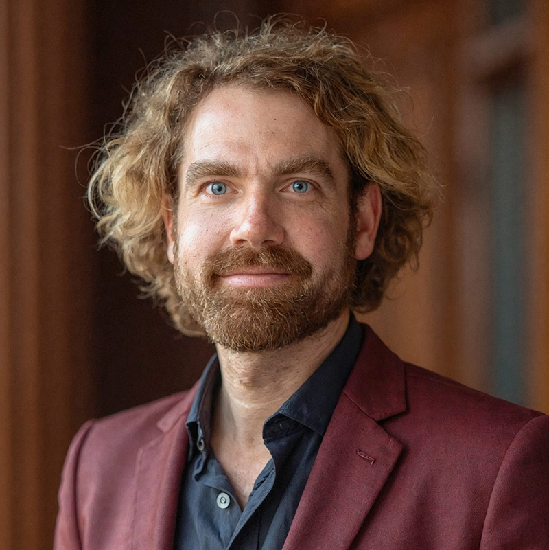 Male presenting person with wavy brown hair, wearing a blue shirt and maroon jacket.